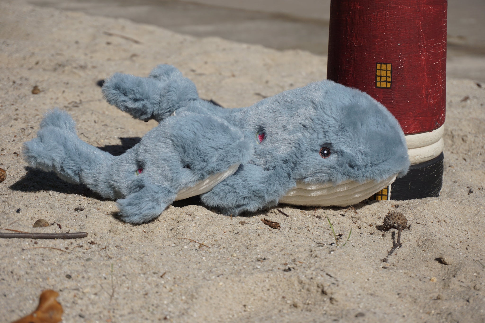 Two gray Mobie Whale Knottie® dog toys sitting on beach next to lighthouse decor piece