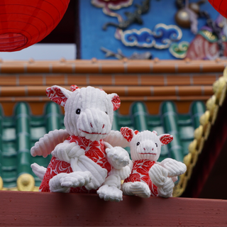 Set of two Lucky dragon plush dog toy with white knotted corduroy limbs, white corduroy head and wings, red and white floral fabric on body, inner ears, and horns, black embroidered eyes, and red embroidered nostrils and mouth in large and small with ornate Asian building in background.