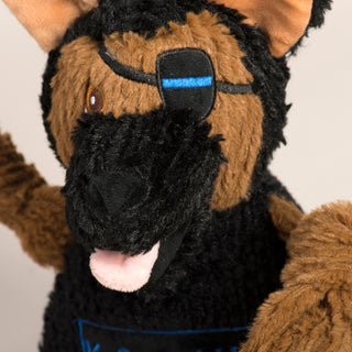 Close up photo of a black and tan German Shepherd dog toy. This is a closeup of his face including his eye patch covering his left eye. On the patch there is a blue line, a tribute to his police work. You can also see his pink tongue and black snout.