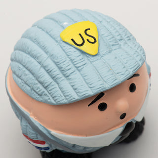 Close up image of ball shaped mail-man dog toy: has blue hat with the letters "US" with light-skin, black eyebrows, black eyes, and black mouth.