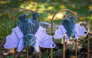 Two Baxter Bat plush toys hanging upside down on a metal fence outdoors, with purple wings and dark grey corduroy bodies.