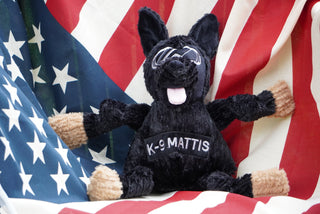 Black shepherd durable plush dog toy. Has black fur, has on a pair of silver goggles, black nose, pink tongue, black knotted limbs, light-brown paws, with the name "K-9 Mattis" across the chest, and is squeaky in front of American flag.
