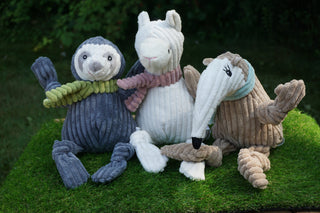 Llama shaped plush dog toy with pink scarf, sitting on grass next to beige furred anteater with light blue scarf, and a gray furred sloth with light green scarf.