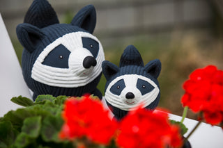 Set of two squeaky ball shaped raccoon dog toy sitting on stone slab: has black and white fur, black tail, white face, black outer-eyes, black nose, and red smile.