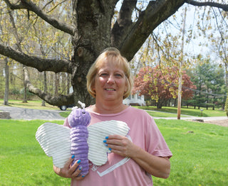 Wendy, the cheerful and detail-savvy financial lead at HuggleGroup, stands smiling in front of a leafy tree, holding a purple butterfly-shaped dog toy—symbolizing her role in ensuring quality and value in every playful product.