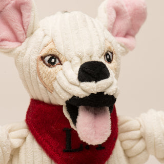 Close up image of dog shaped durable plush corduroy dog toy to show detail in face: has white fur, pink inner-ear, beige outer-ear, black eyes, white pupils, plush-out black nose, black mouth, pink tongue, with red bandana that has the initials "L.T", and is squeaky with knotted limbs.