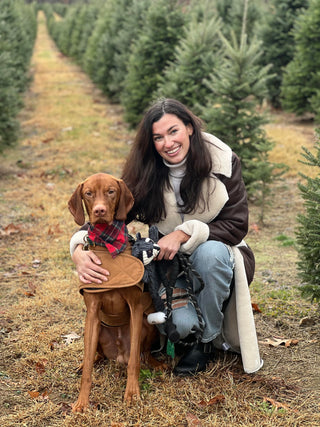 Image of Campbell Brady crounched down next to her brown furred dog sitting down with trees in the background.