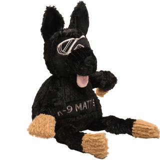 Side view of black shepherd shaped durable plush dog toy. Has black fur, has on a pair of silver goggles, black nose, pink tongue, black knotted limbs, light-brown paws, with the name "K-9 Mattis" across the chest, and is squeaky.