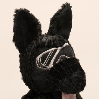 Close up image of dog shaped durable plush dog toy, with black fur, silver goggles, black nose, and pink tongue.