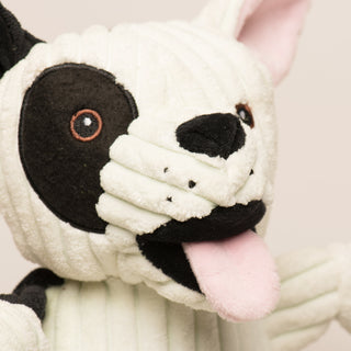 Close up image of a Jonny Justice durable plush corduroy dog toy to show details in face: The dog has black and white fur, right outer-eye is black, has brown eyes, white pupils, plush out black nose, black mouth, pink tongue, and is squeaky.