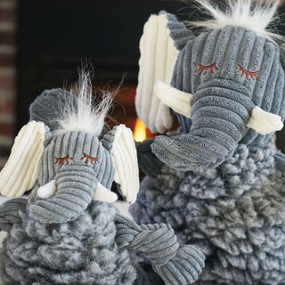 Set of two elephant shaped durable plush dog toys: grey fluffy HuggleFleece® body, white hair, white inner-ear, white tusks, and is squeaky with knotted limbs sitting in front of fireplace.