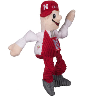 Side view of University of Nebraska Lil Red mascot plush dog toy with beige, skin tone face and hands, white long sleeved undershirt, red corduroy overalls with embroidered university logo, grey corduroy shoes, big nose, embroidered eyes and mouth, and red hat with words "LIL' RED" and blond hair poking out underneath hat.