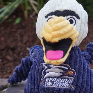 Close up of Georgia Southern University Gus the eagle Shaped plush dog toy: has white head, black unibrow, white eyes, black pupils, yellow beak, black mouth with a red tongue, the body arms and legs are navy blue, navy blue shirt has the university logo on it, has yellow feet, and is squeaky with knotted limbs.