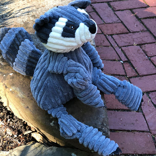 Left side view of raccoon shaped plush dog toy sitting on a stone slab looking up.