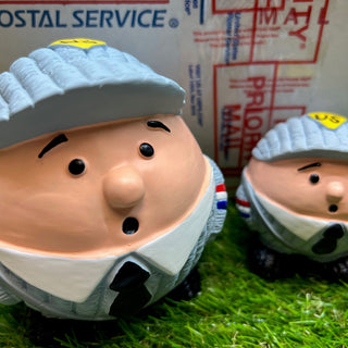 Set of two squeaky latex ball shaped mail carrier dog toys: has blue hat, black eyebrows, black eyes, black mouth, blue uniform, black tie and shoes in front of mailing envelope.