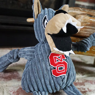 Close up view of North Carolina State University Mr. Wuf mascot durable corduroy plush dog toy with grey body, head, and knotted limbs, tan muzzle and inner ears, wide open black mouth with pointy white teeth, embroidered black and white eyes, and embroidered university logo on chest.