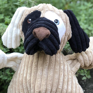 Portrait view of dog shaped plush dog toy: has brown fur, with white furred right ear, black furred left ear, black fur on right outer-eye, white fur on left outer-eye, black lips, brown nose, and white right arm.