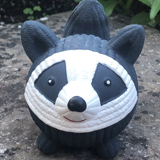 Squeaky ball shaped raccoon dog toy sitting on stone slab: has black and white fur, black tail, white face, black outer-eyes, black nose, and red smile.