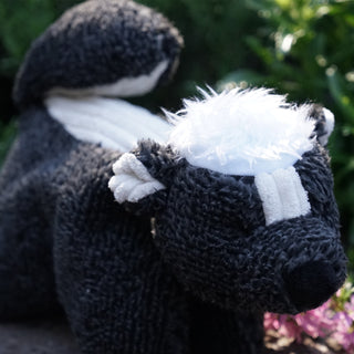 Side view of skunk shaped durable Squooshie™ plush dog toy with fuzzy black and gray body, white corduroy inner-ears and streak down back, fluffy white hair on head, and black eyes and nose.