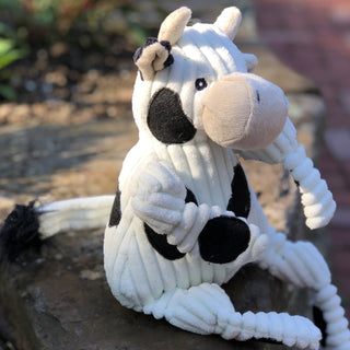 Side view of cow shaped durable plush corduroy dog toy with white fur with black spots, light beige horns, black eyes, white pupils, black nose, light beige mouth, and is squeaky with knotted limbs.