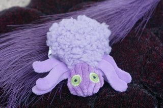 Light purple spider cat toy with bright green embroidered eyes.  the spider is sitting on a darker purple background that looks like velvet and brighter purple feathers.  