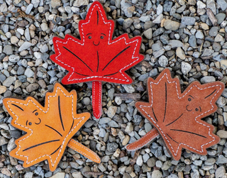 Set of three leaf-shaped all-natural leather dog chew toys with black eyes, smile, and details, and white stitching: red, burnt orange, and brown sitting on gravel.
