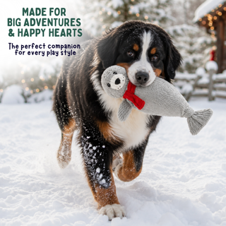 A Bernese mountain dog running through the snow while carrying a gray knitted fish-shaped plush toy with a red bow tie. Text on the top left reads 'MADE FOR BIG ADVENTURES & HAPPY HEARTS' and 'The perfect companion for every play style.