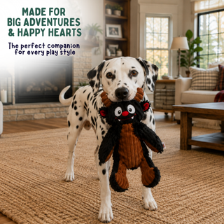 A Dalmatian stands in a cozy living room holding a plush monster toy in its mouth. The toy is black with brown sections, red eyes, and small red ears.