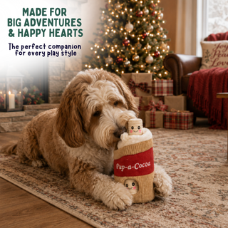 A fluffy light brown dog lies on a patterned rug in a cozy living room, chewing on a plush cup-shaped toy labeled 'Pup-a-Cocoa.' A decorated Christmas tree, wrapped gifts, fireplace, and sofa are visible in the background.