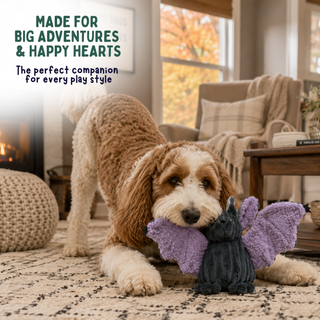 A fluffy brown and cream dog lies on a patterned rug in a cozy living room, holding a small purple plush bat toy in its mouth. The text on the top left reads, 'MADE FOR BIG ADVENTURES & HAPPY HEARTS The perfect companion for every play style.