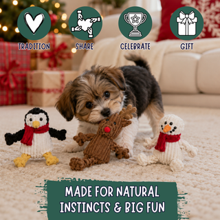 A small puppy stands on a carpeted floor in a living room setting, surrounded by three knitted holiday-themed dog toys: a penguin, a reindeer, and a snowman. Decorative wrapped gifts, a sofa, and festive lights are blurred in the background.