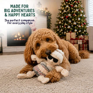 A fluffy golden-brown dog lies on a beige carpet in a cozy living room, holding a plush puppy toy. A Christmas tree, fireplace, and wrapped gifts are visible in the background.