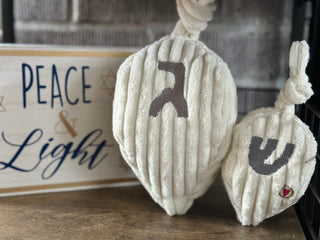 Two white dreidel plush dog toys displayed on a shelf next to a 'Peace & Light' Hanukkah sign.
