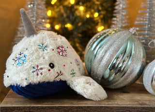 Durable plush narwhal dog toy with white faux-fur with shiny metallic rainbow snowflake design, gray horn, blue corduroy belly, and a knotted tail sitting next to Christmas ornament decorations.