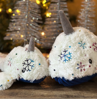 Set of two narwhal-shaped durable plush dog toys in large and small: both have white faux-fur with shiny metallic rainbow snowflake design, gray horn, blue corduroy belly, and a knotted tail with silver Christmas trees in background.