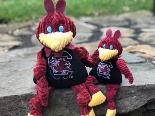 Set of two University of South Carolina, Cocky mascot durable plush corduroy dog toys in large and small with burgundy head, burgundy knotted wings and legs, blue eyes, yellow beak and feet, black shirt with university logo across the chest.