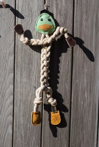 Green duck all natural rope dog toy with a green leather face, yellow leather bill, black leather hair, brown leather hands and tan leather feet, with a rope body, arms, and legs hanging on wall.
