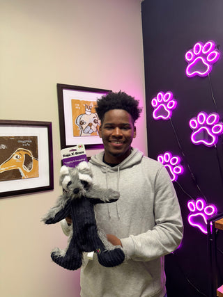 Image of Jose Sosa standing while holding a Raccoon shaped plush and durable dog toy. in front of a wall with lights and paintings. 