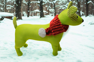 Green knitted dog with a red scarf standing in a snowy forest