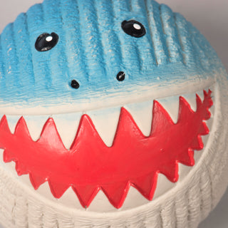 Close up of squeaky ball shaped shark dog toy to show corduroy texture on latex: with blue skin, black eyes, white pupils, black nose, red mouth, white teeth, and a white stomach.