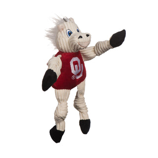 Left view of University of Oklahoma Sooner plush dog toy: has white fur, white hair, blue eyes, white pupils, white furred plush-out nose and lips, black nose, beige lips, has on a red shirt with university logo, black hands and feet, and knotted limbs.
