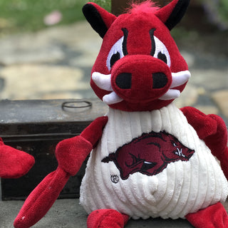 Close up of University of Arkansas Tusk the Razorback red boar mascot durable plush corduroy dog toy with knotted limbs wearing white shirt and University of Arkansas logo on the front.