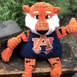 Auburn University Aubie the tiger mascot durable plush corduroy dog toy with white fluffy face, knotted limbs wearing navy blue shirt and Auburn University logo on the front sitting on bench.