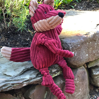 Left view of burgundy furred fox plush dog toy, sitting on a stone slab.