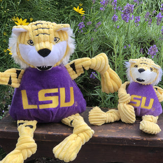 Set of two LSU Mike the Tiger mascot plush corduroy dog toy with yellow knotted limbs, yellow head, fluffy white hair around face, white inner ears, black stripes on head and limbs, white face, gold embroidered eyes, black nose, black open mouth with white fangs, wearing purple shirt with gold embroidered LSU logo across chest sitting on bench in front of plants.