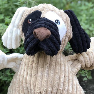Portrait view of dog shaped plush dog toy: has brown fur, with white furred right ear, black furred left ear, black fur on right outer-eye, white fur on left outer-eye, black lips, brown nose, and white right arm. 