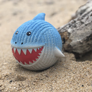 Lifestyle image of squeaky ball shaped shark dog toy on beach: has blue skin, black eyes, white pupils, black nose, red mouth, white teeth, and a white stomach.