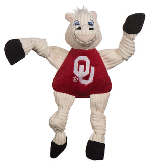 University of Oklahoma Sooner plush dog toy: has white fur, white hair, blue eyes, white pupils, white furred plush-out nose and lips, black nose, beige lips, has on a red shirt with university logo, black hands and feet, and knotted limbs. 