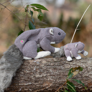 Lifestyle image of large and small gray furred squirrel plush dog toys on a tree log. 